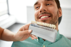 man smiles in dental exam chair while dental professional holds teeth shades to his mouth and man asks does professional teeth whitening cause sensitivity