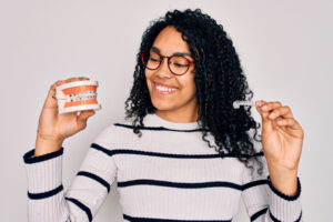 a woman wearing a stripe sweater and glasses holds mouth models that have braces on them and an invisalign