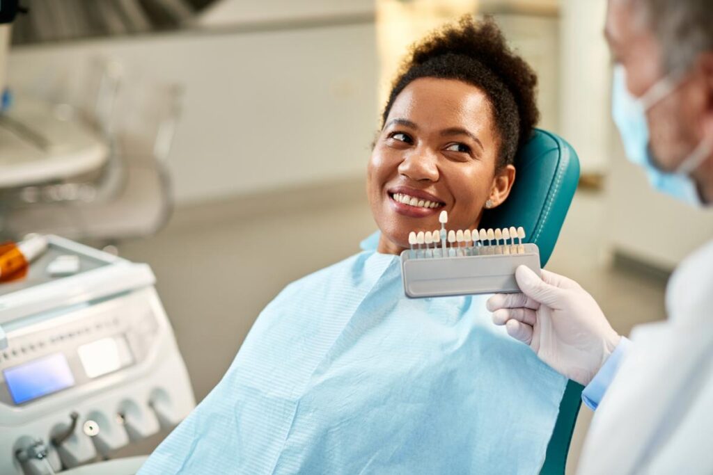 a woman patient sits in a chair smiling while her dentist holds up shades of porcelain veneers to match her other teeth